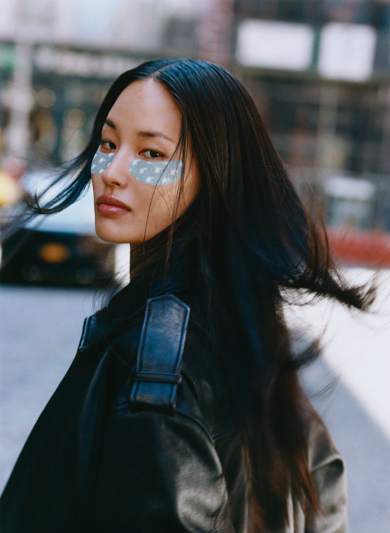 Portrait of a woman with eye patches, wearing a black leather jacket in an urban setting, with blurred buildings in the background.