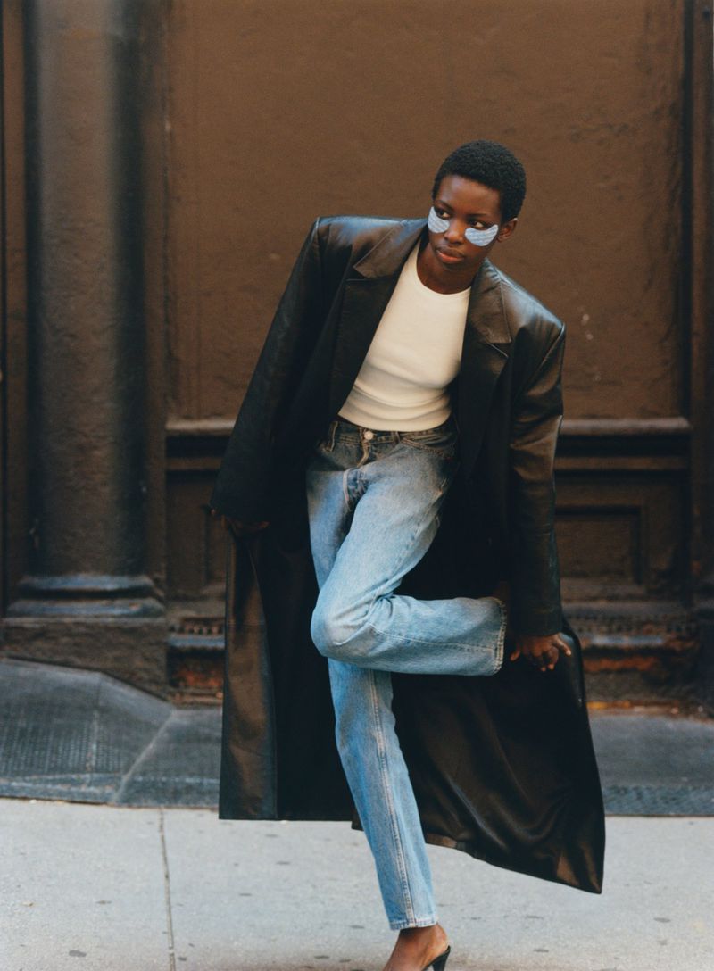 Fashionable adult female poses outdoors in a black trench coat, top, and jeans, complemented by stylish blue under-eye patches.