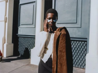Model Abuk Manyang Adeer wearing under-eye patches, a cream knit cardigan, and a brown suede jacket against a building exterior.