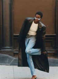 Model wearing under-eye patches, a white t-shirt, blue jeans, and a long black leather coat outdoors against a building.