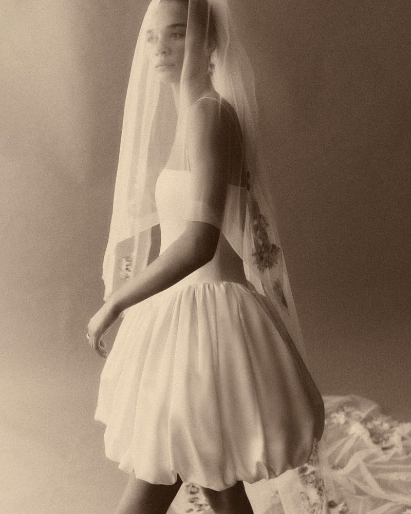 A woman poses in a short, white dress and sheer, floral veil. The soft lighting and warm tones give the image a vintage aesthetic.