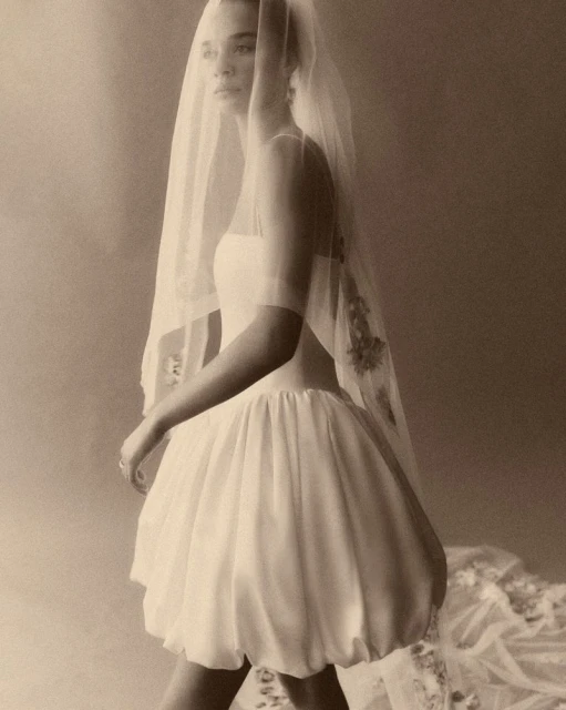 A woman poses in a short, white dress and sheer, floral veil. The soft lighting and warm tones give the image a vintage aesthetic.