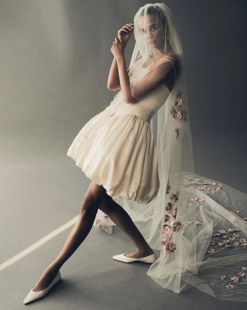 Model poses in a short, light-colored dress and long floral-accented veil against a grey backdrop in a fashion editorial setting.