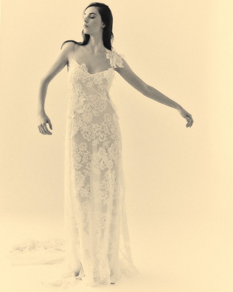 Elegant woman in a lace dress with an asymmetric neckline poses in a bright studio with arms outstretched against a neutral backdrop.