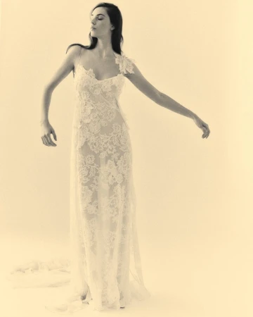 Elegant woman in a lace dress with an asymmetric neckline poses in a bright studio with arms outstretched against a neutral backdrop.