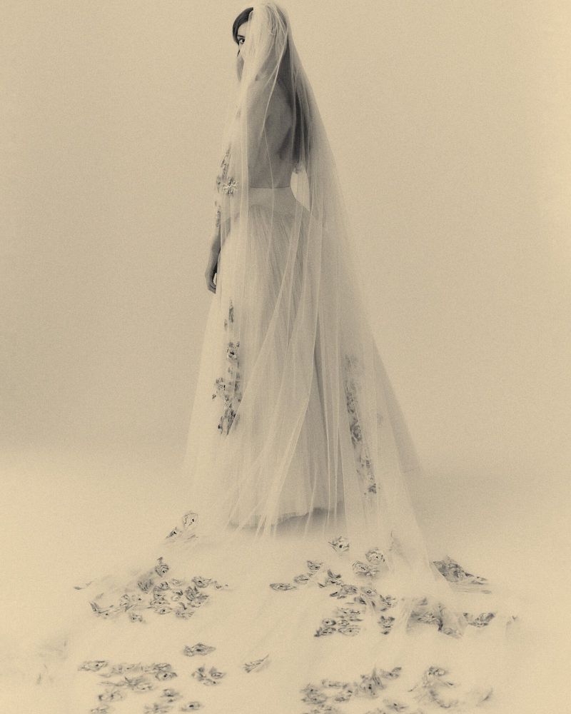 A woman poses in a light-toned photo studio wearing a long white wedding dress with a floral-embroidered veil trailing behind her.