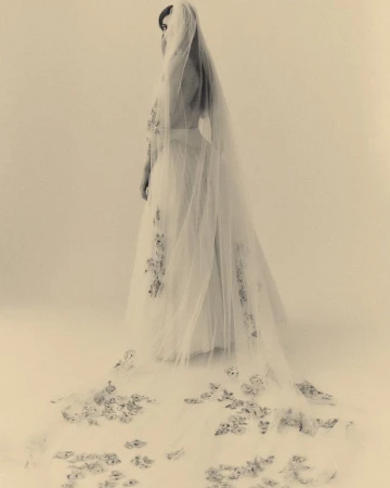 A woman poses in a light-toned photo studio wearing a long white wedding dress with a floral-embroidered veil trailing behind her.