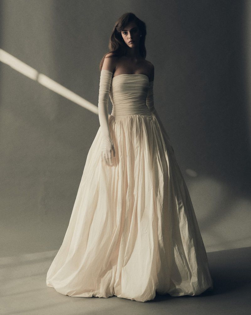 A woman poses in a strapless white gown and long gloves, with studio lighting and a grey background. Fashion editorial shot.