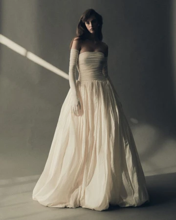 A woman poses in a strapless white gown and long gloves, with studio lighting and a grey background. Fashion editorial shot.