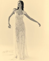 Model wearing a delicate sheer white floral lace gown with one embellished strap, posing against a warm off-white studio backdrop.