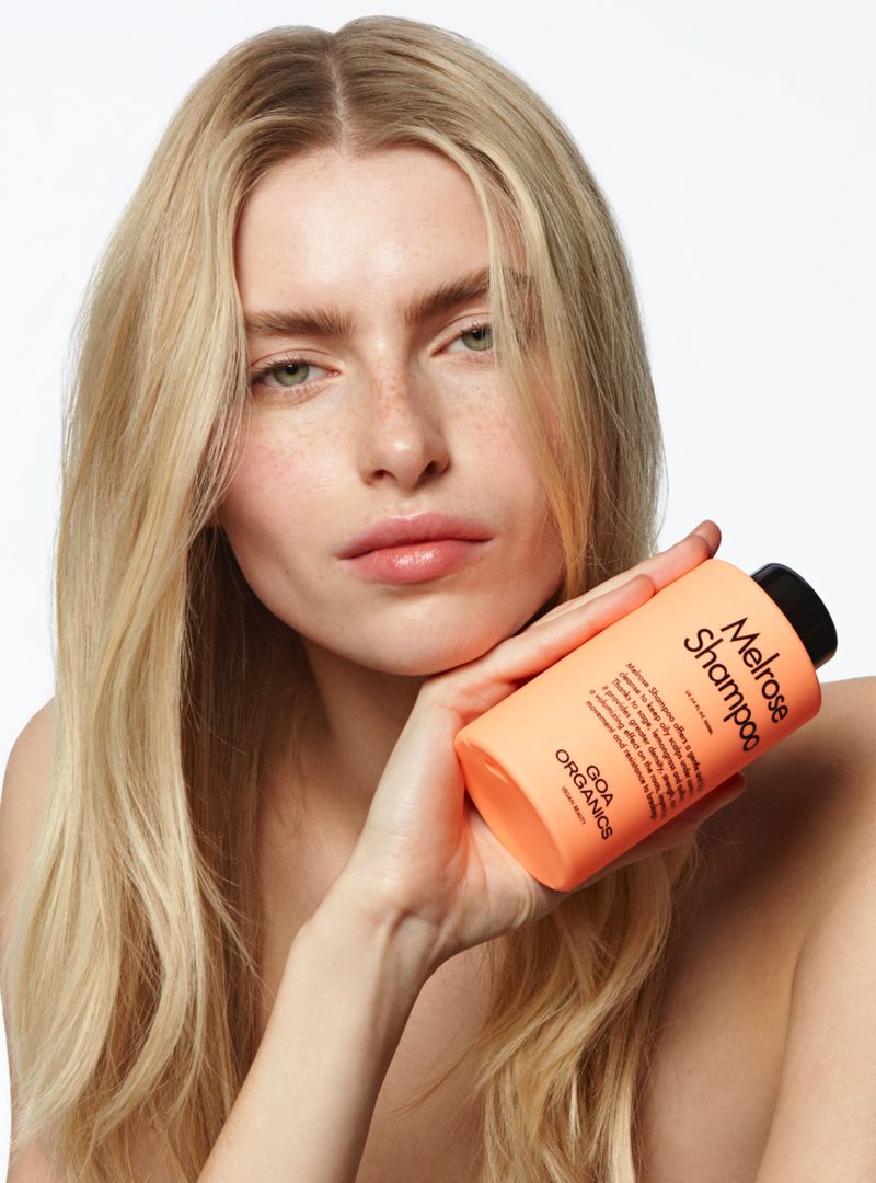 A woman with long blonde hair holds an apricot-colored bottle of Melrose Shampoo in a close-up studio portrait against a white backdrop.