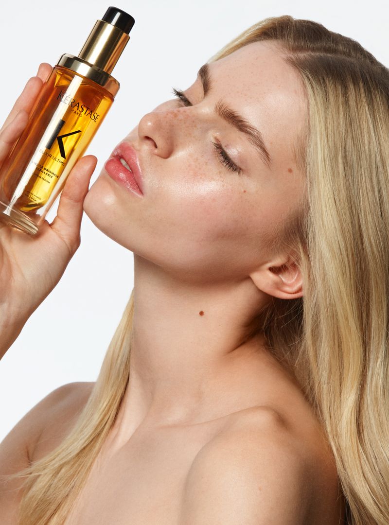 Close-up of a blonde-haired woman holding a bottle of Kérastase hair oil against a clean, bright background in a beauty editorial style.