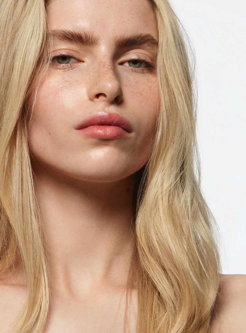 Close-up portrait of a young fair-skinned woman with blonde wavy hair, subtle makeup, and freckles, against a bright white background.