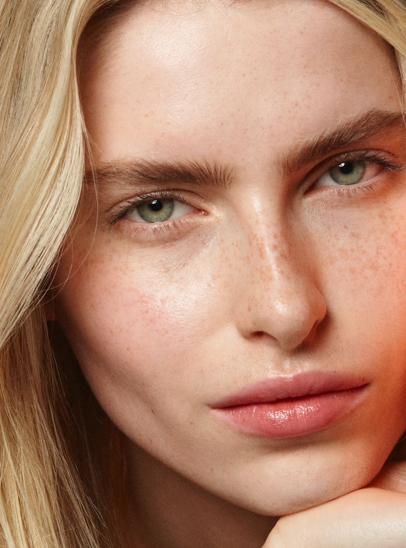 Close-up portrait of a young fair-skinned woman with light blonde wavy hair and freckles, looking directly at the camera indoors.