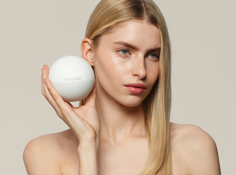 Woman with bare shoulders holding a white spherical container of Cocunat beauty product against a neutral background in a beauty product shoot.