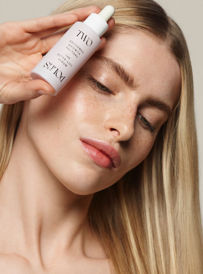 Fair-skinned blonde woman holds a skincare serum bottle with a dropper, posing against a neutral background, beauty product shot.