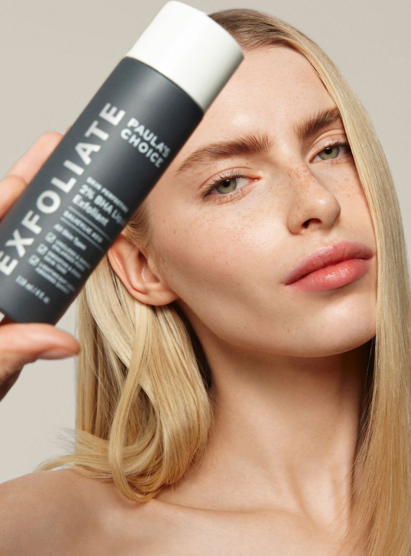 Blonde woman posing with Paula's Choice EXFOLIATE product in a beauty shot, neutral background, clear skin, close-up product placement.