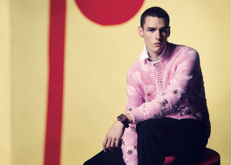 Man in a pink sweater sitting against a yellow backdrop with a red vertical line and circle. Fashion editorial shot. Indoor studio.