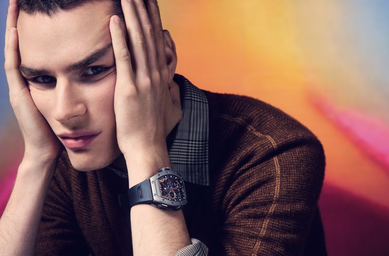 Man poses with hands on face wearing brown plaid shirt and wristwatch against colorful background in a studio setting.