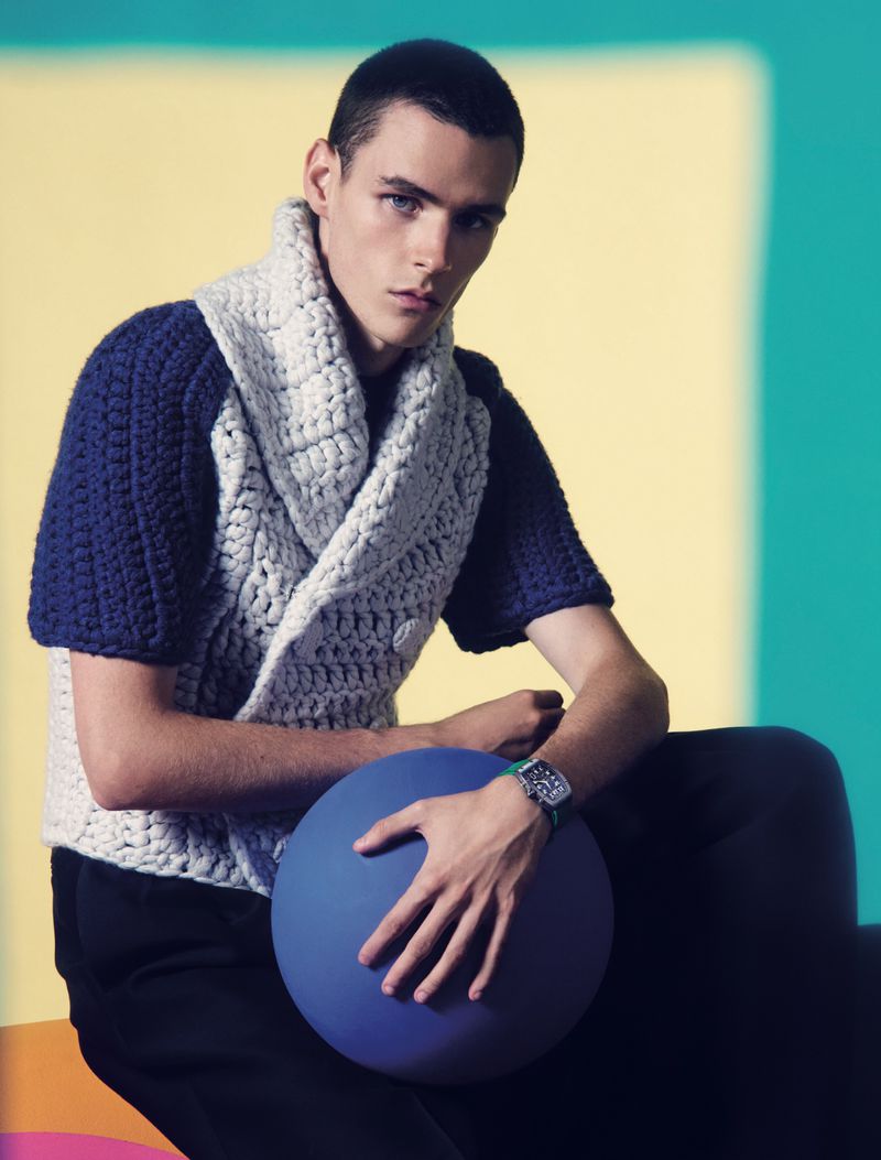 Portrait of a young adult male in a knit sweater, holding a blue ball against a colorful background of yellow, teal, and orange.