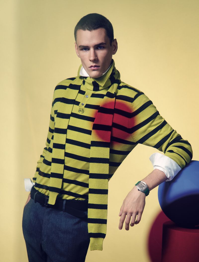 A young adult man poses indoors with a yellow-and-black striped shirt, navy pants, watch, and colored spheres on a yellow background.