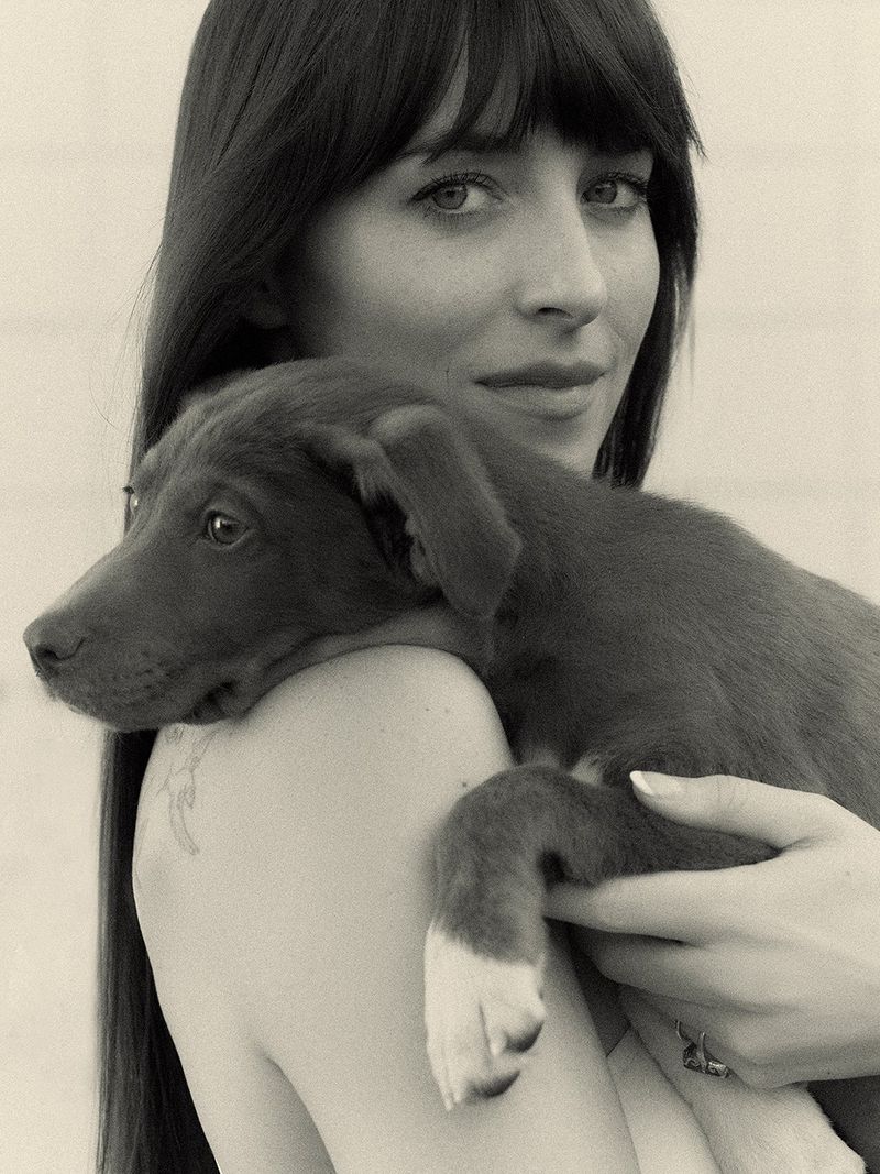 Monochrome portrait of a woman with long, dark hair, gazing forward while cradling a brown puppy with white paws on her shoulder.