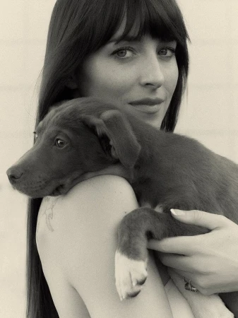 Monochrome portrait of a woman with long, dark hair, gazing forward while cradling a brown puppy with white paws on her shoulder.