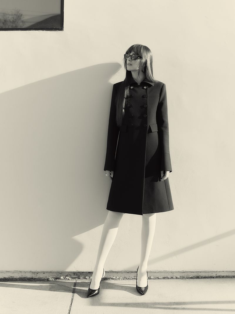 Monochrome fashion shot of a slender woman in a dark coat, white stockings, and heels posing against a white wall with shadow.