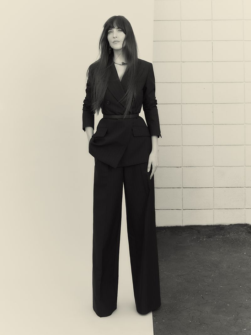 A woman in a dark suit poses in front of a two-tone tiled wall, creating a stylish and professional image with a vintage aesthetic.