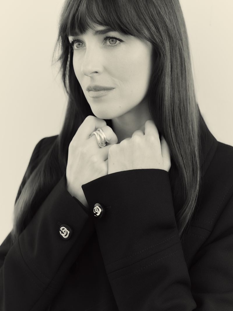 Close-up portrait of a brunette woman with bangs wearing a black jacket and silver rings, against a neutral studio backdrop.