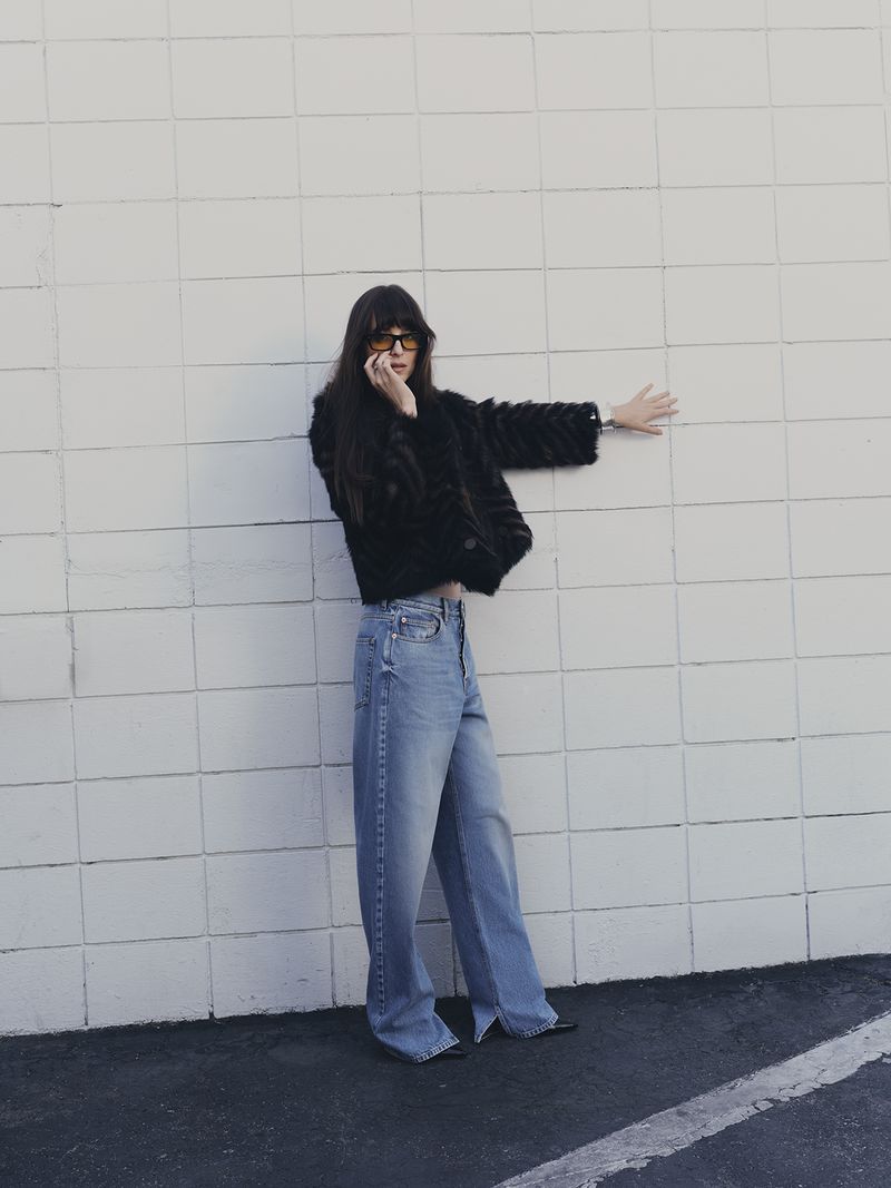 A stylish woman with sunglasses and a fur jacket poses against a white wall. She's wearing jeans and has her hand up against the wall.