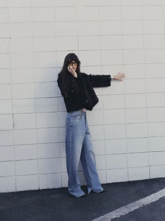 A stylish woman with sunglasses and a fur jacket poses against a white wall. She's wearing jeans and has her hand up against the wall.