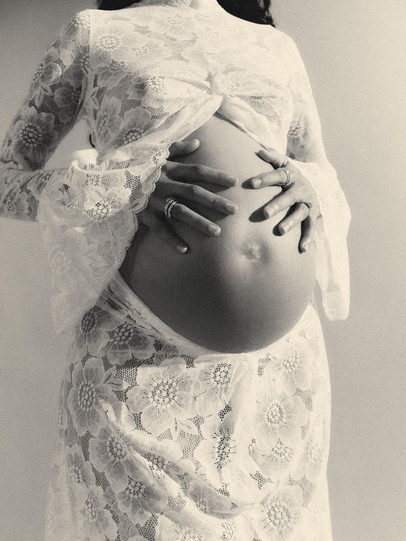 Pregnant woman in a white lace dress poses indoors, hands resting on her belly, monochrome aesthetic.
