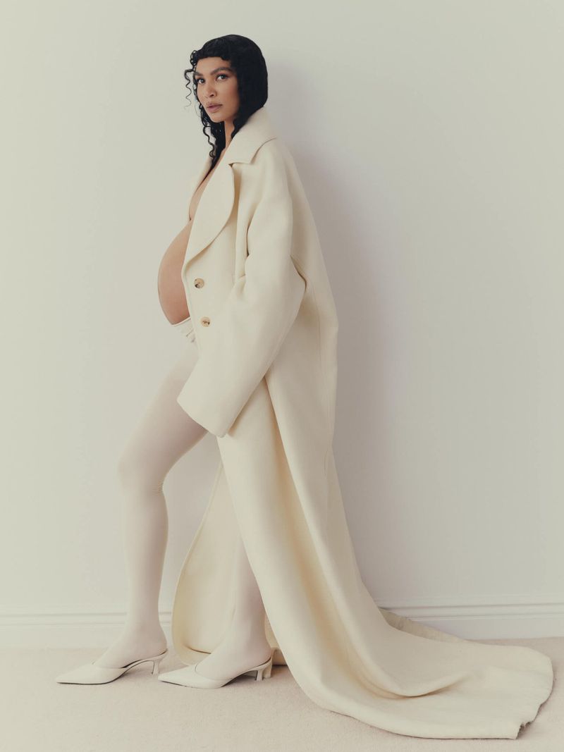 Pregnant woman in white coat posing in a studio, showcasing maternity fashion and elegant style against a neutral background.