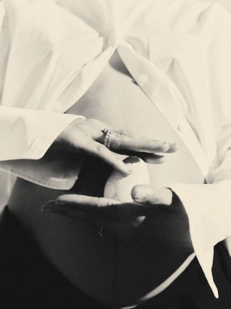 Monochrome close-up of a pregnant woman's belly with hands holding an egg, wearing a white shirt with studio lighting.