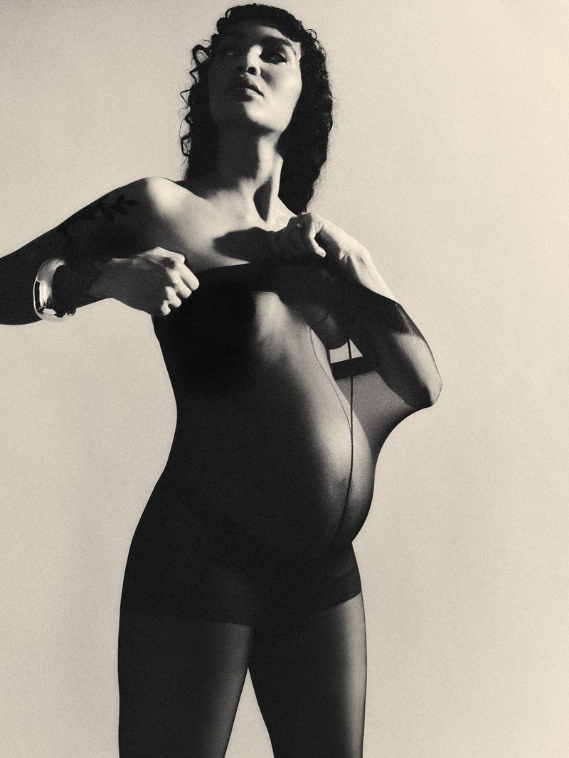 Pregnant woman in sheer tights poses in a close-up studio portrait with neutral lighting and simple background, showing pregnancy fashion.