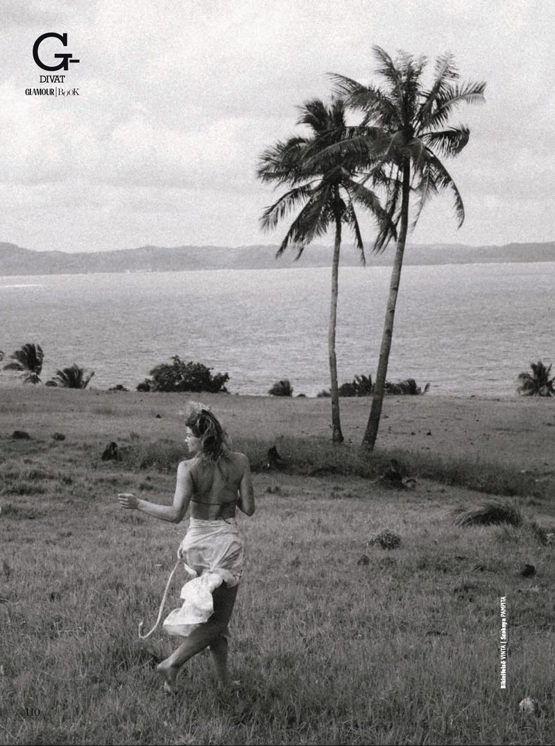 Woman in bikini and sarong walks on grassy hill with palm trees near the ocean; monochrome lifestyle editorial fashion photography.