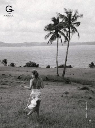 Woman in bikini and sarong walks on grassy hill with palm trees near the ocean; monochrome lifestyle editorial fashion photography.