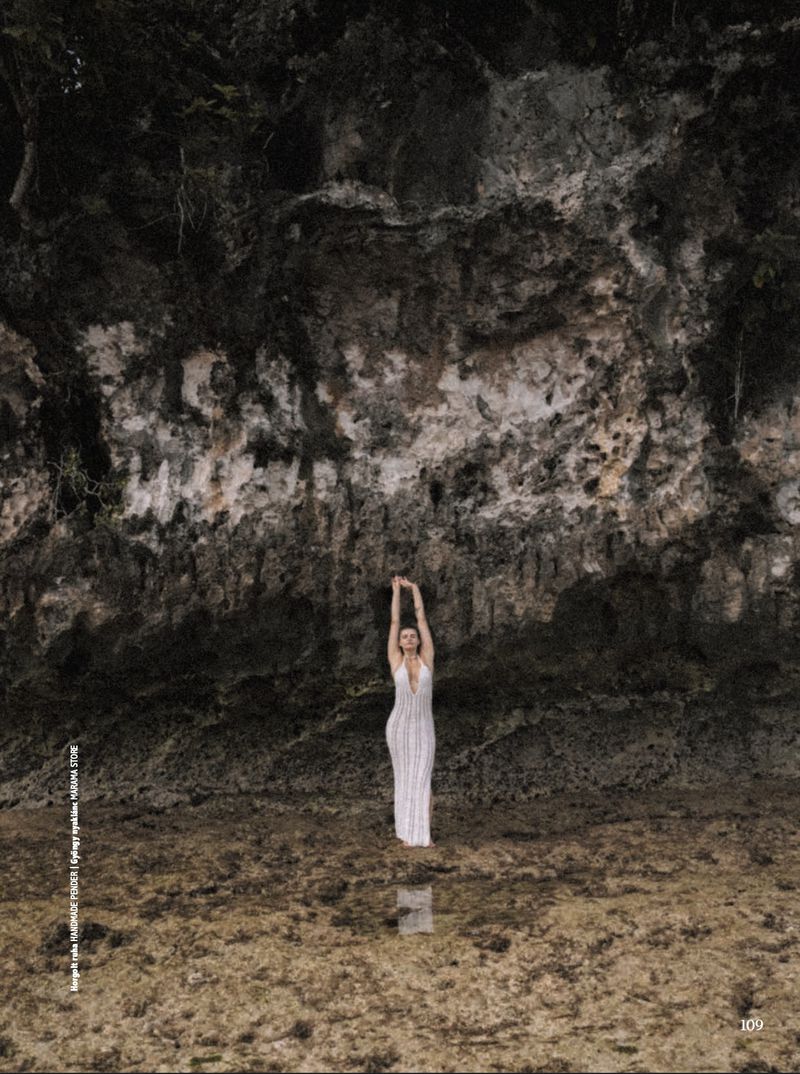 A woman in a white dress stands with her arms raised in front of a rocky cliff face, creating a serene and eye-level portrait.