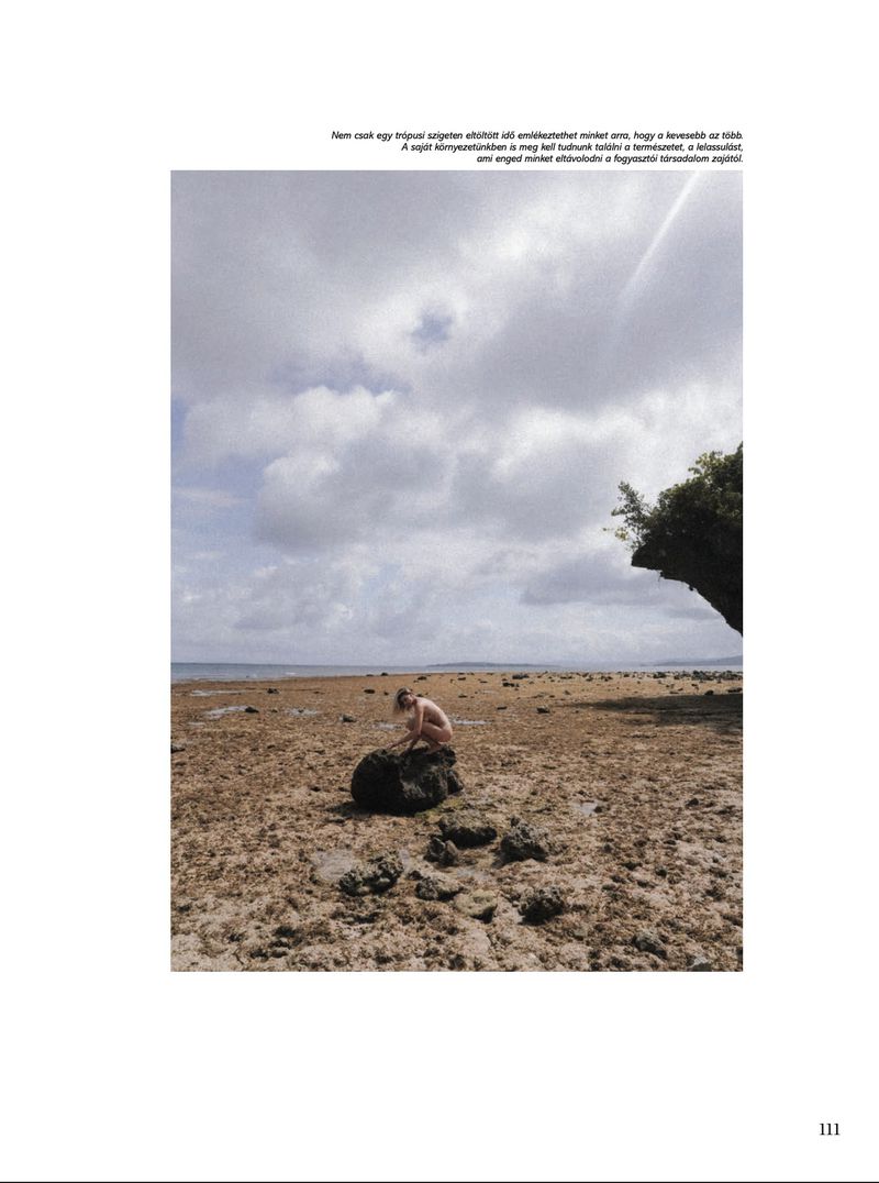 Person sits on a rock at a sandy beach under a cloudy sky. The horizon line and the landscape are visible, creating a tranquil seaside scene.