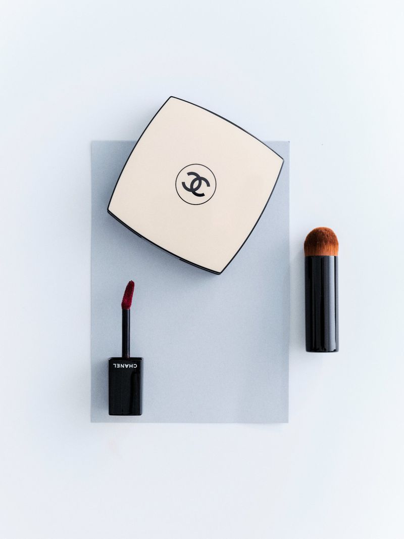 Overhead shot of Chanel makeup: a cream square compact with the logo, a black lip gloss applicator, and a black makeup brush.