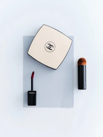 Overhead shot of Chanel makeup: a cream square compact with the logo, a black lip gloss applicator, and a black makeup brush.