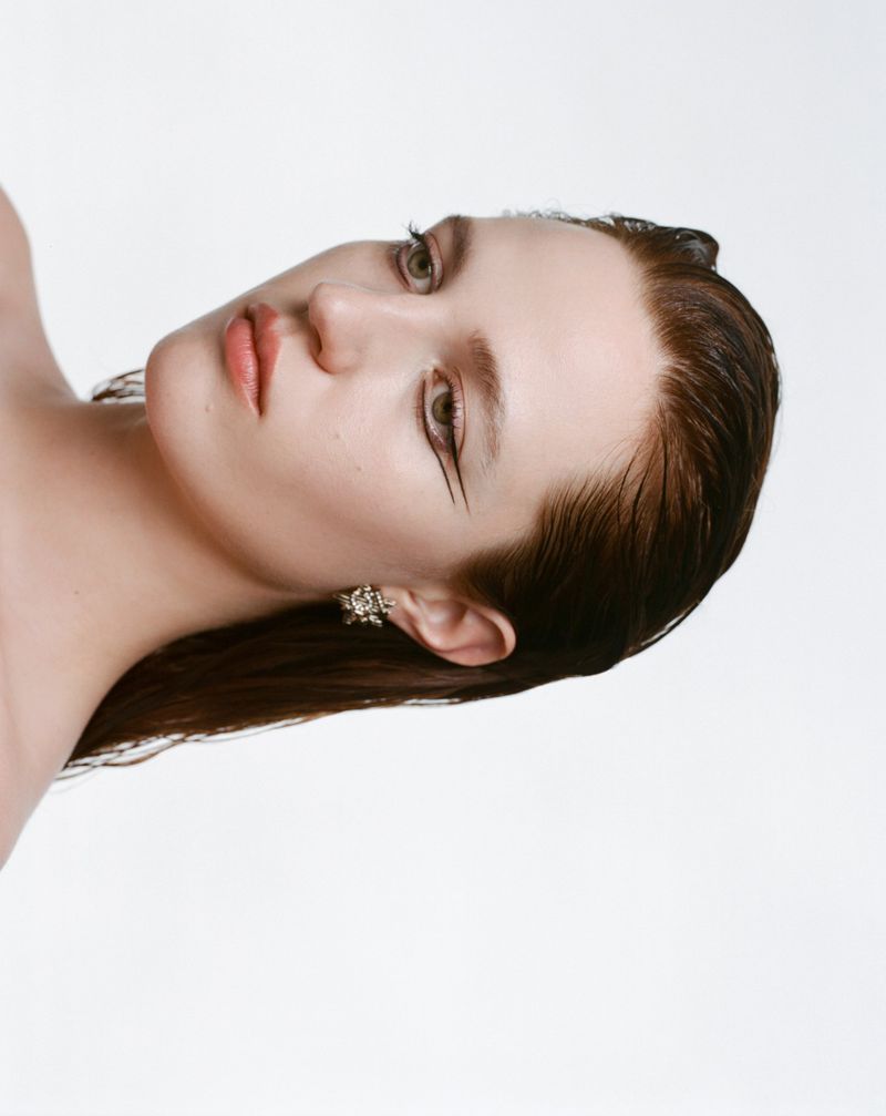 Close-up beauty shot of a young adult woman with wet brunette hair, defined eyeliner, and statement floral earrings against a white background.