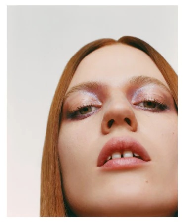 Close-up shot of a young woman with light skin, red hair, and striking violet eyeshadow, viewed from a low angle against a light backdrop.