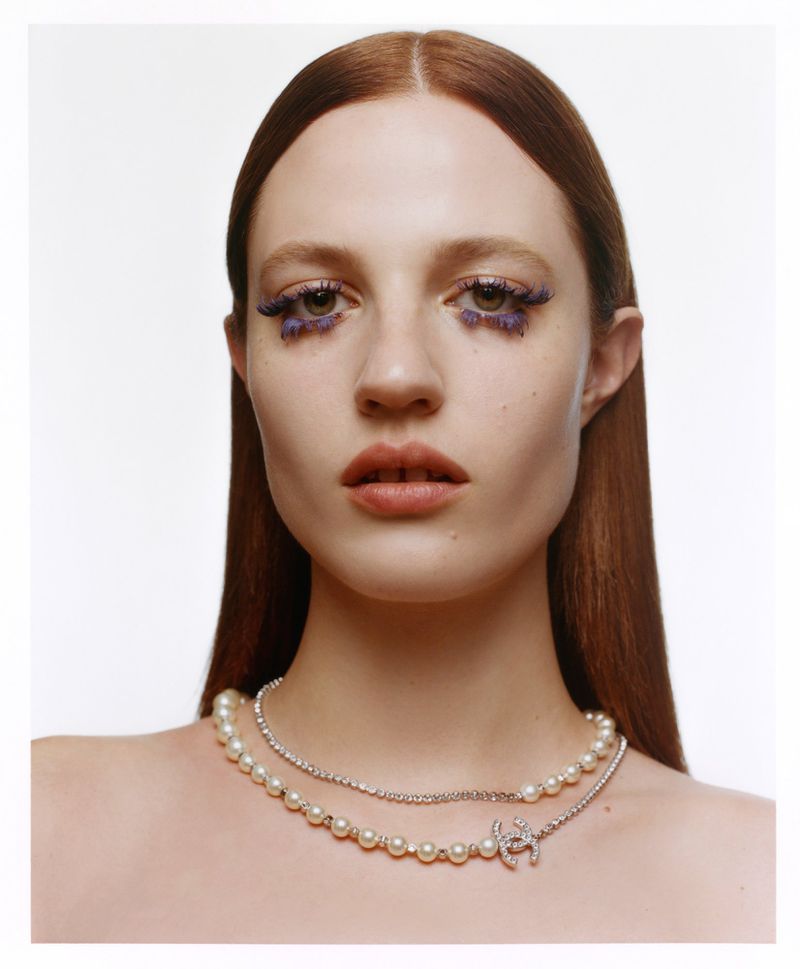 Close-up portrait of a woman with auburn hair, pearl and diamond jewelry, and dramatic violet eye makeup against a bright white background.