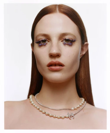 Close-up portrait of a woman with auburn hair, pearl and diamond jewelry, and dramatic violet eye makeup against a bright white background.
