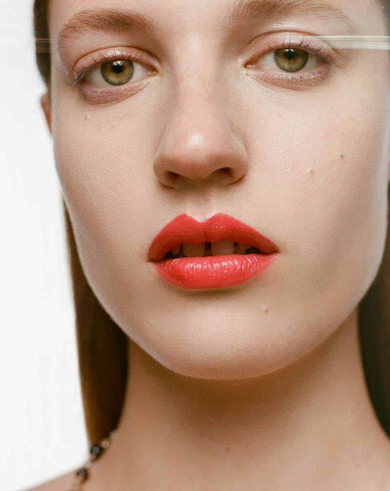 Close-up portrait of a woman with light skin, green eyes, and red lips. The woman is wearing a necklace and has a neutral expression.
