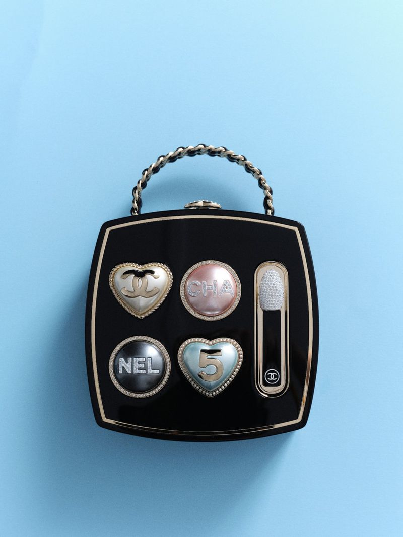 Overhead shot of a black Chanel compact with a silver chain handle and round and heart shaped decorative buttons on a light blue background.