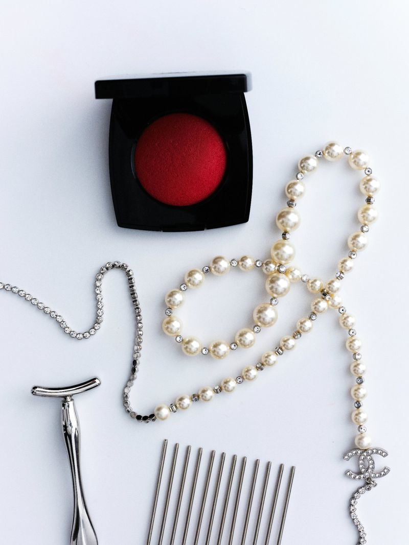 Flat lay of red blush, pearl necklace, metal comb, silver razor, and crystal chain on a clean white surface.