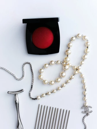 Flat lay of red blush, pearl necklace, metal comb, silver razor, and crystal chain on a clean white surface.